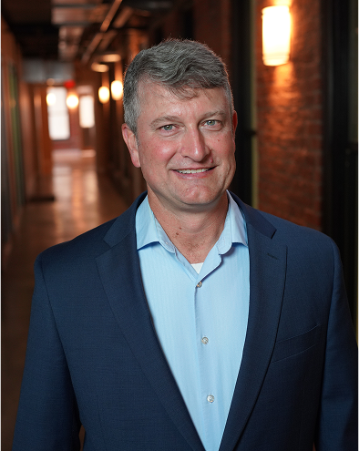 Man in a blue suit smiling in a dimly lit hallway.