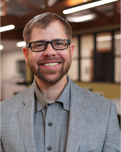 Smiling man with glasses and beard in a gray suit in an office.