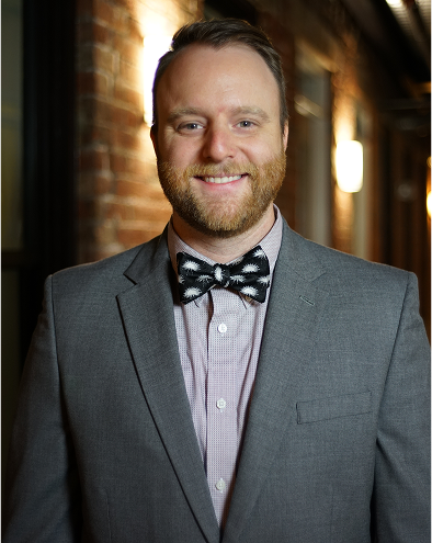 Man in a gray suit with a polka dot bow tie, smiling in a hallway.