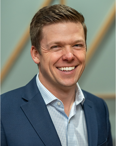 Smiling man in a blue suit, light shirt, neutral background.