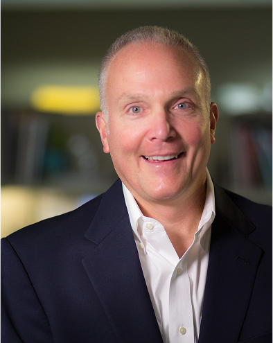 Smiling man in a suit with blurred bookshelves in the background.