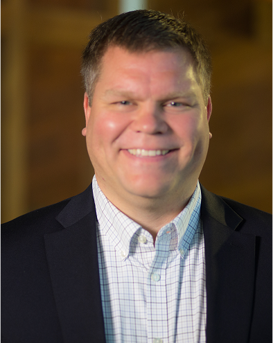 Smiling man in a suit and checkered shirt, blurred background.
