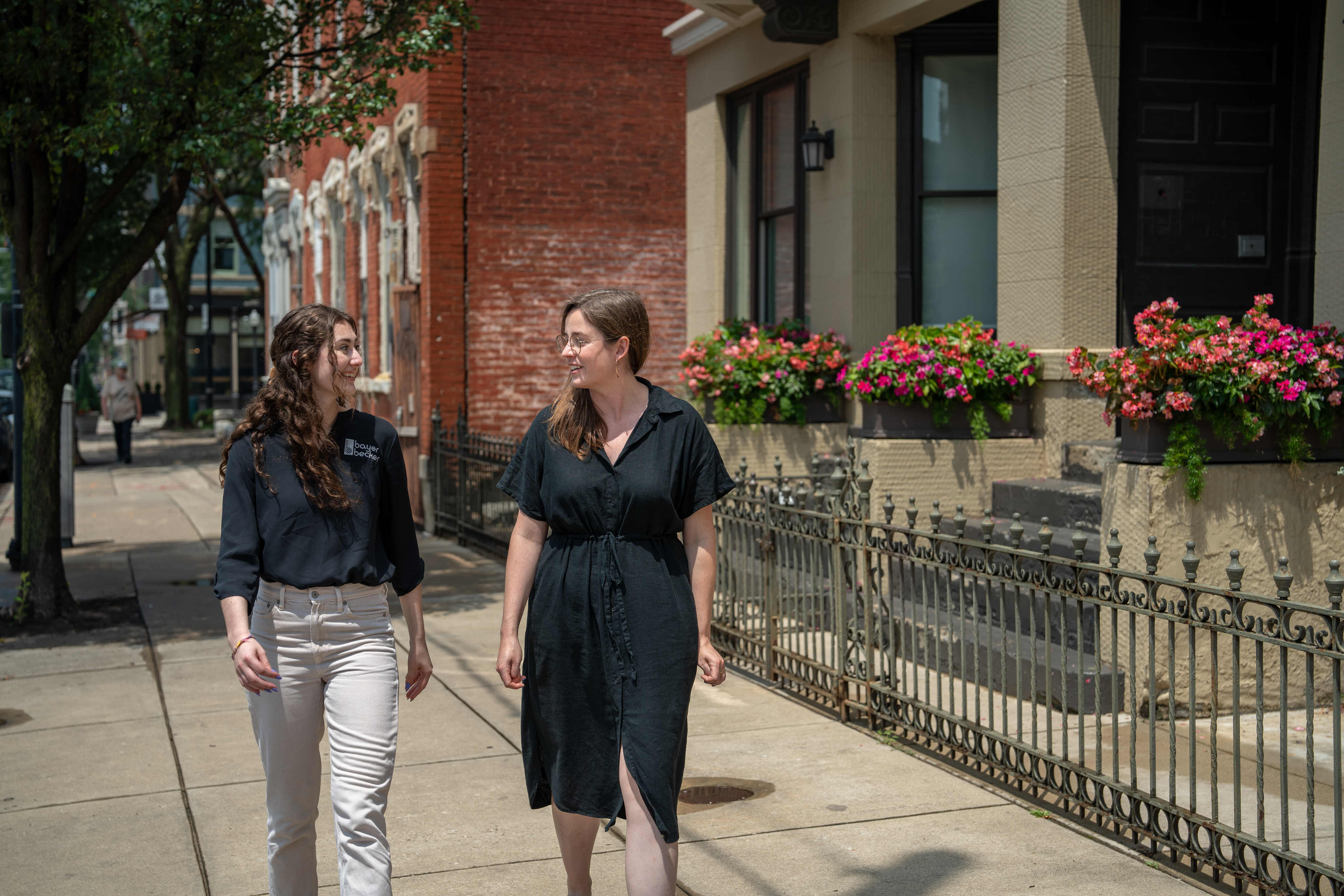 Two women walking on a sidewalk near a brick building with flowers.