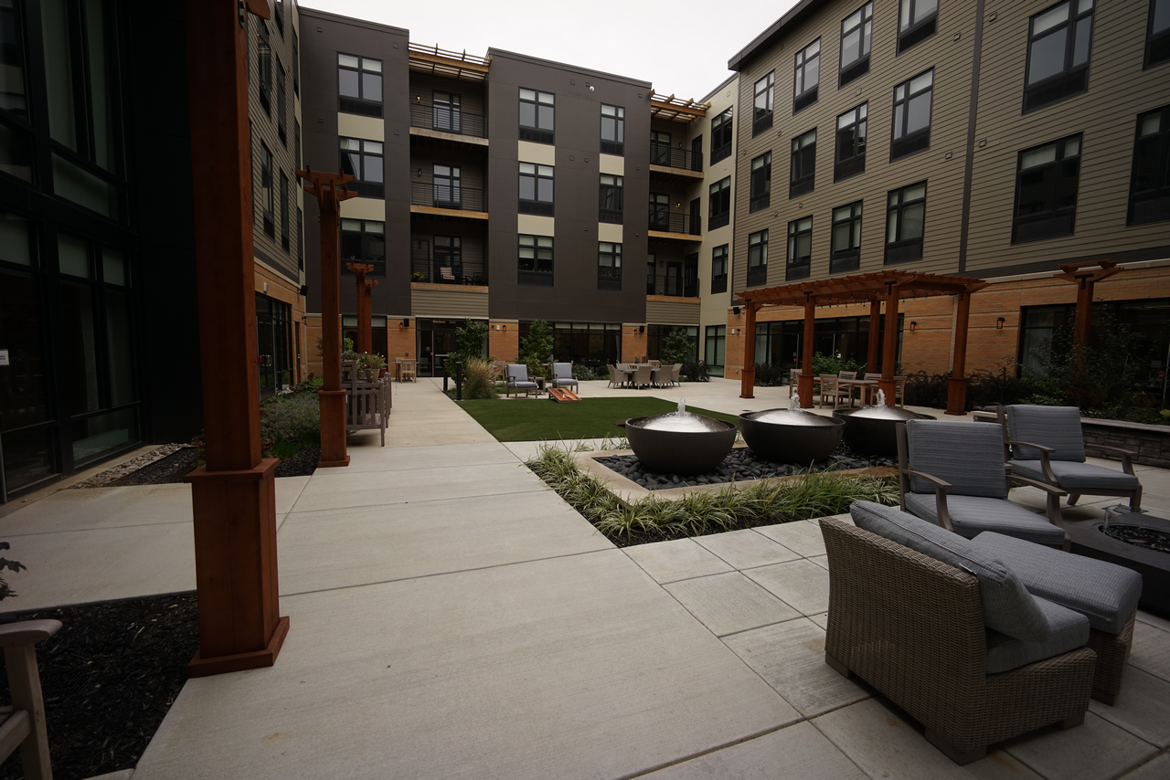 Courtyard with seating, umbrella tables, and greenery surrounded by multi-story buildings.