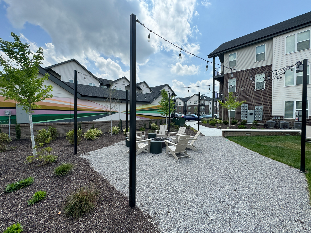 Outdoor seating area with chairs, strung lights, and modern buildings under a partly cloudy sky.