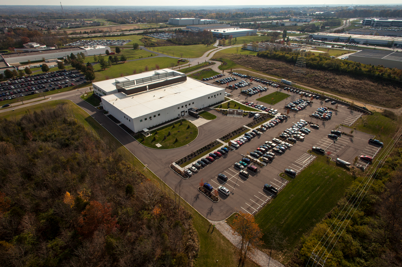 Aerial view of a large white building with parking lot, surrounded by fields and trees.