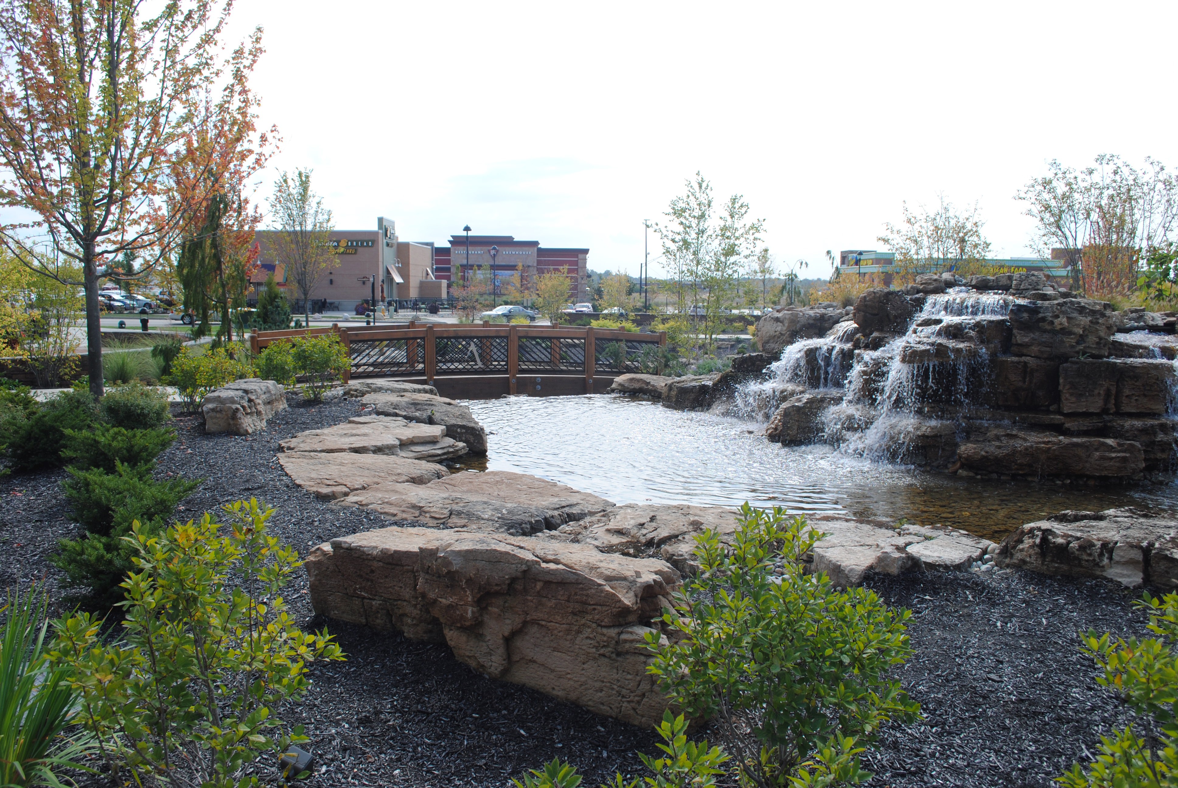 Waterfall flows into a pond surrounded by rocks and greenery in an urban park setting.