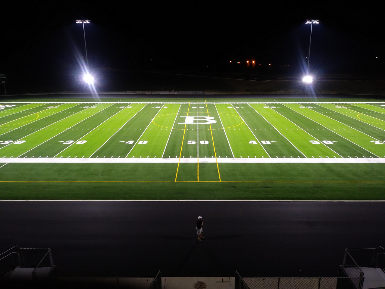 Floodlit football field at night, one person standing on track.
