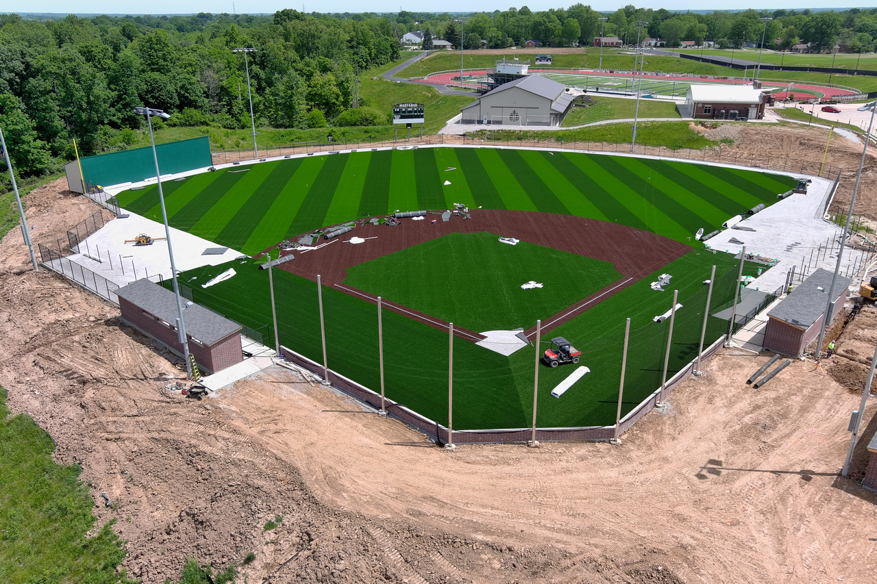 Baseball field with green and brown turf, surrounded by trees and buildings.
