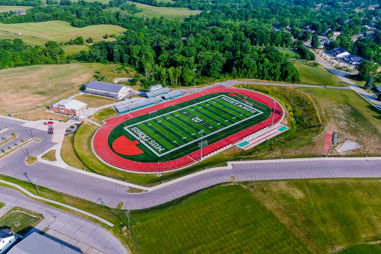 Aerial view of a football field with a red track, surrounded by greenery and roads.