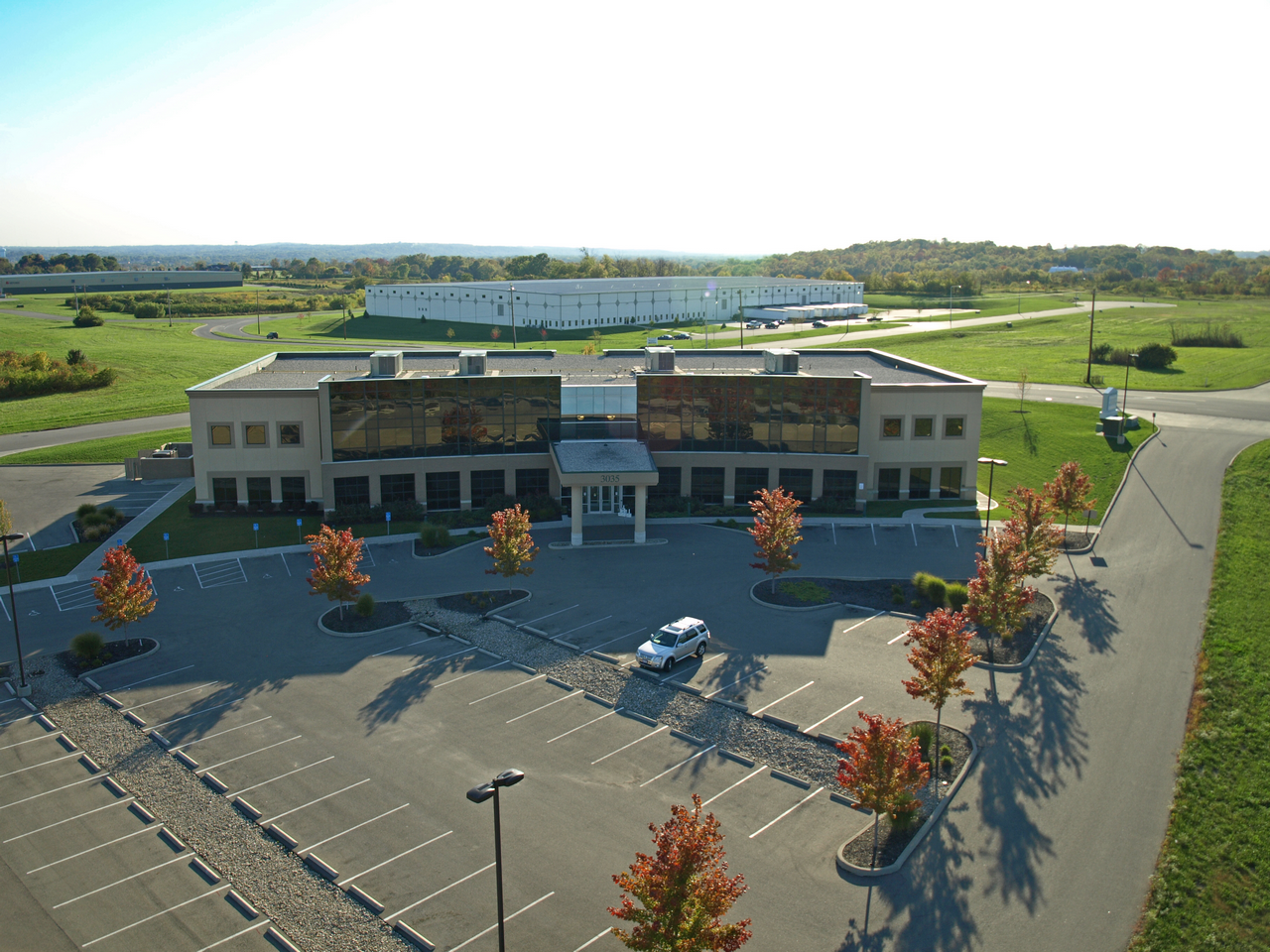 Modern office building with empty parking lot, surrounded by green fields and autumn trees.