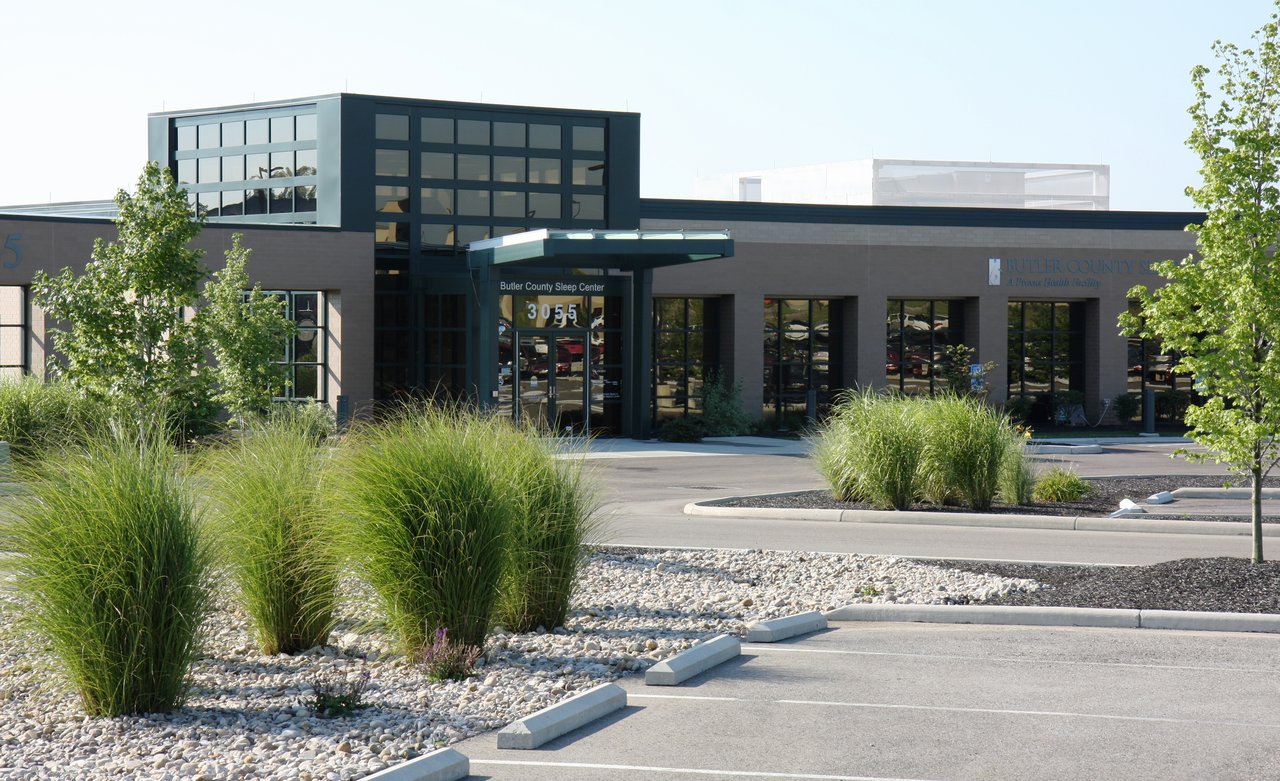 Modern building with glass entrance, surrounded by greenery and parking area.
