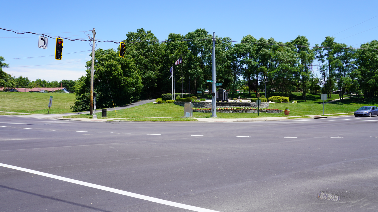 Intersection with traffic lights, trees, and a grassy park area under a clear blue sky.