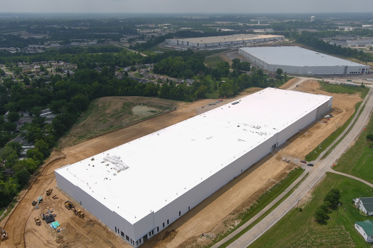 Large white warehouse under construction in a suburban area, surrounded by trees and roads.