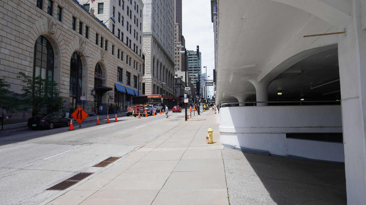 City street with buildings, sidewalk, orange cones, and parked cars.