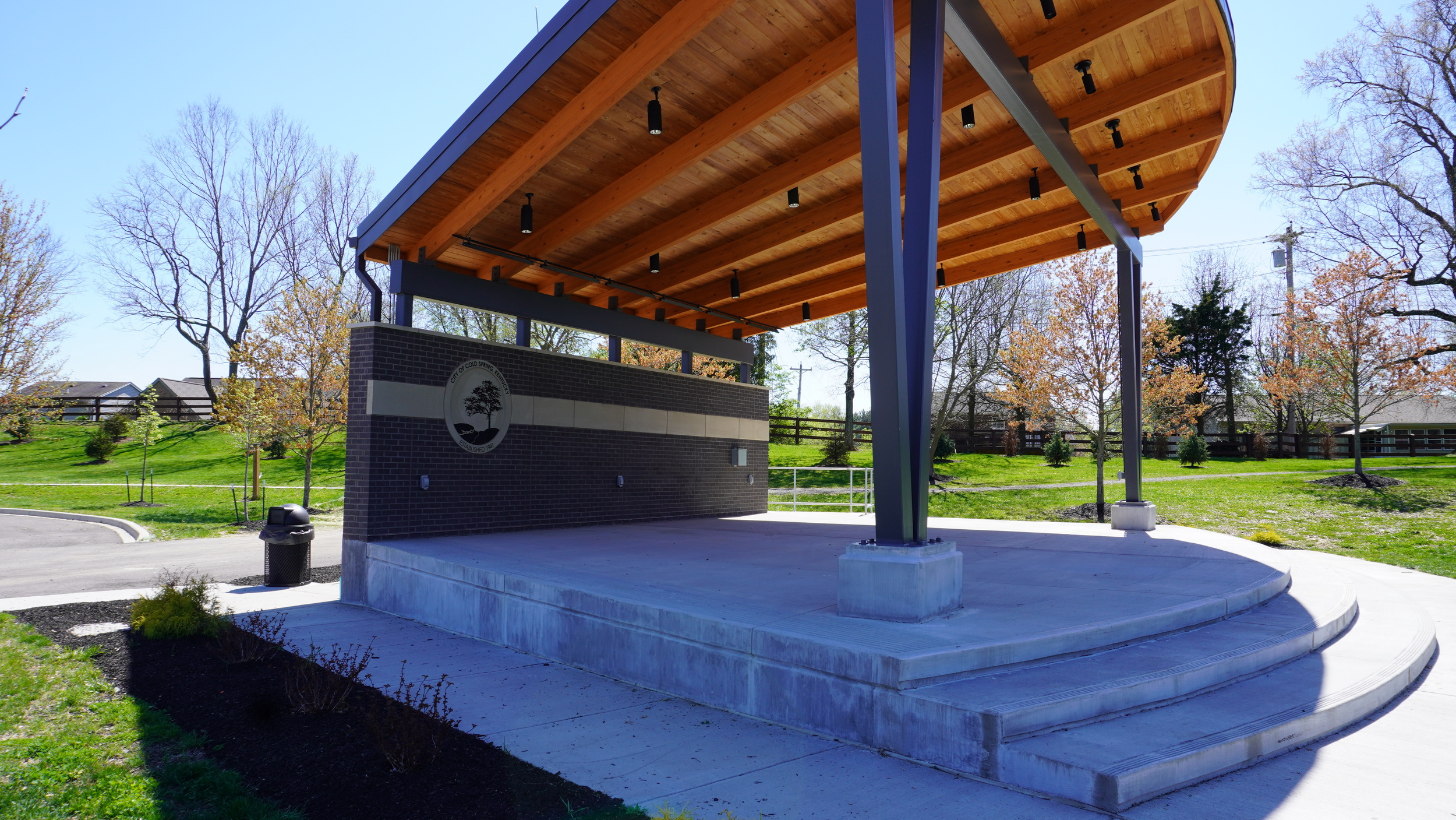 Outdoor pavilion with wooden roof and concrete stage. Trees and grass in background.
