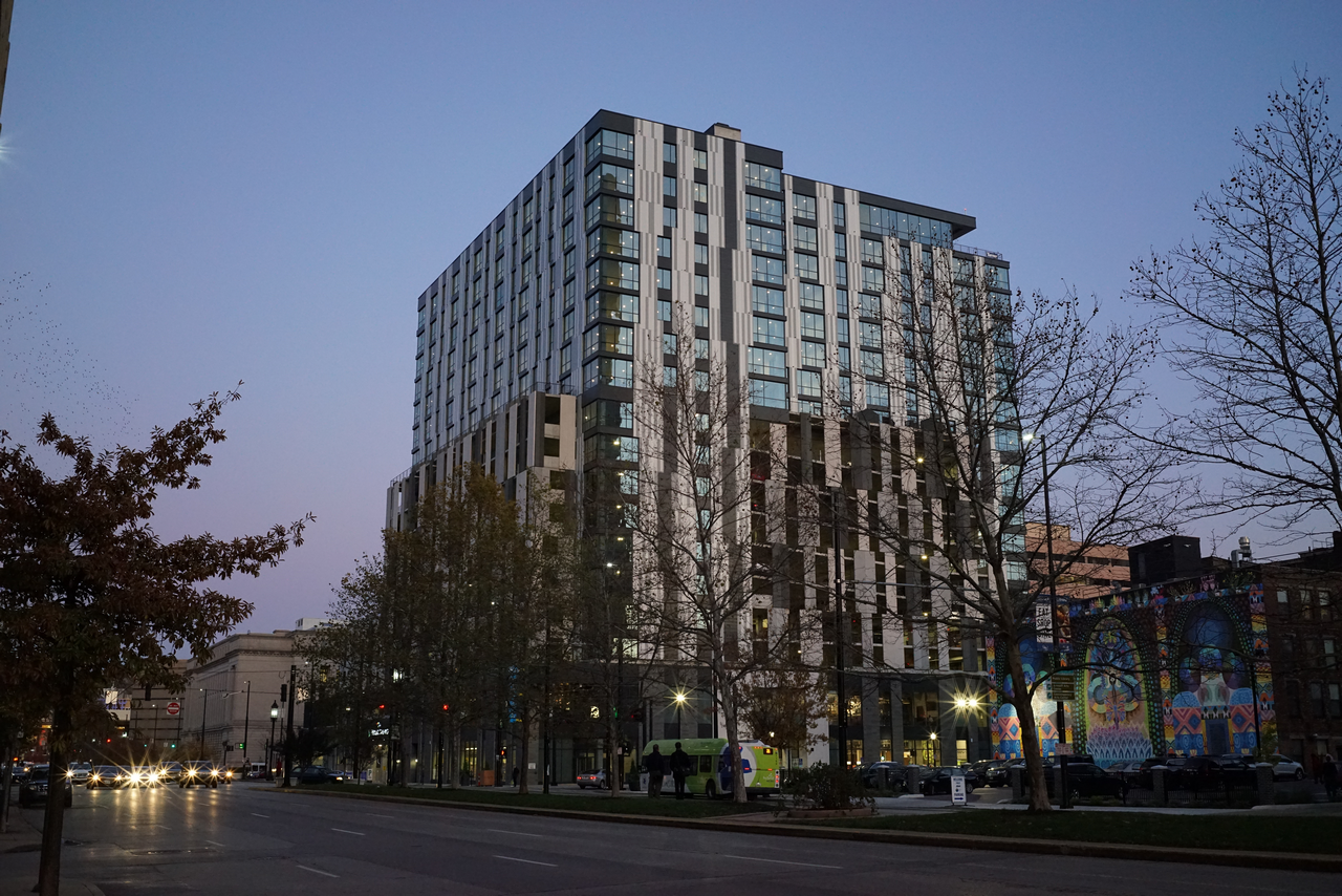 Modern high-rise building at dusk, streetlights illuminate trees and graffiti.