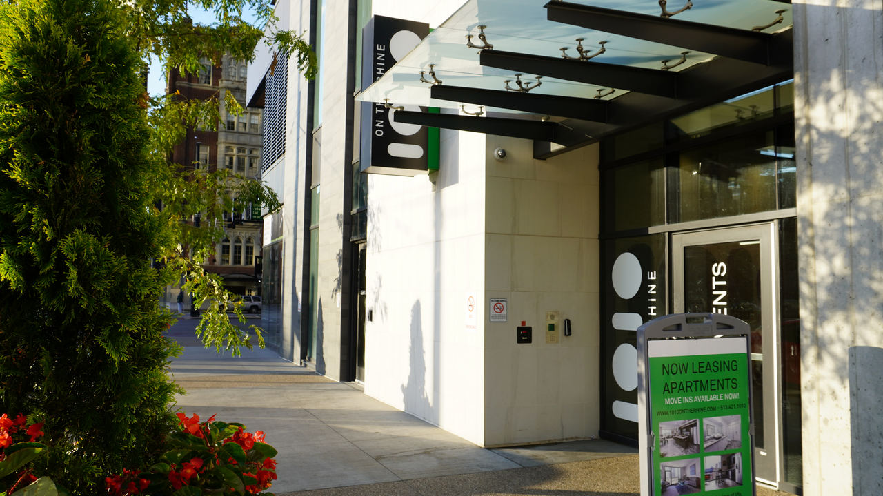 Modern building entrance with glass canopy and greenery against a city street backdrop.