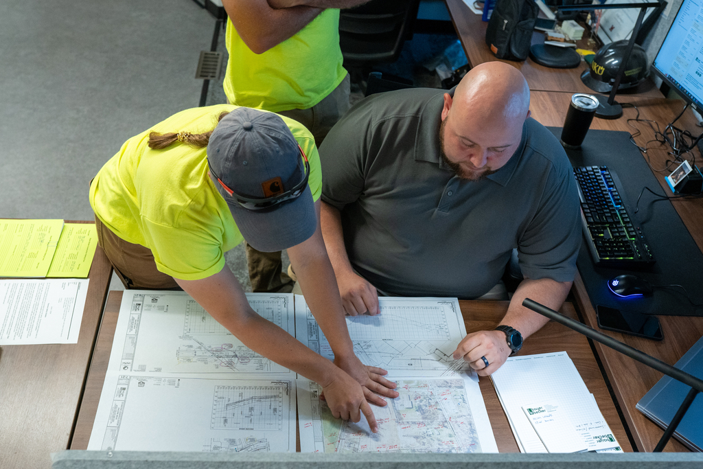 Two people reviewing blueprints at a desk in an office.