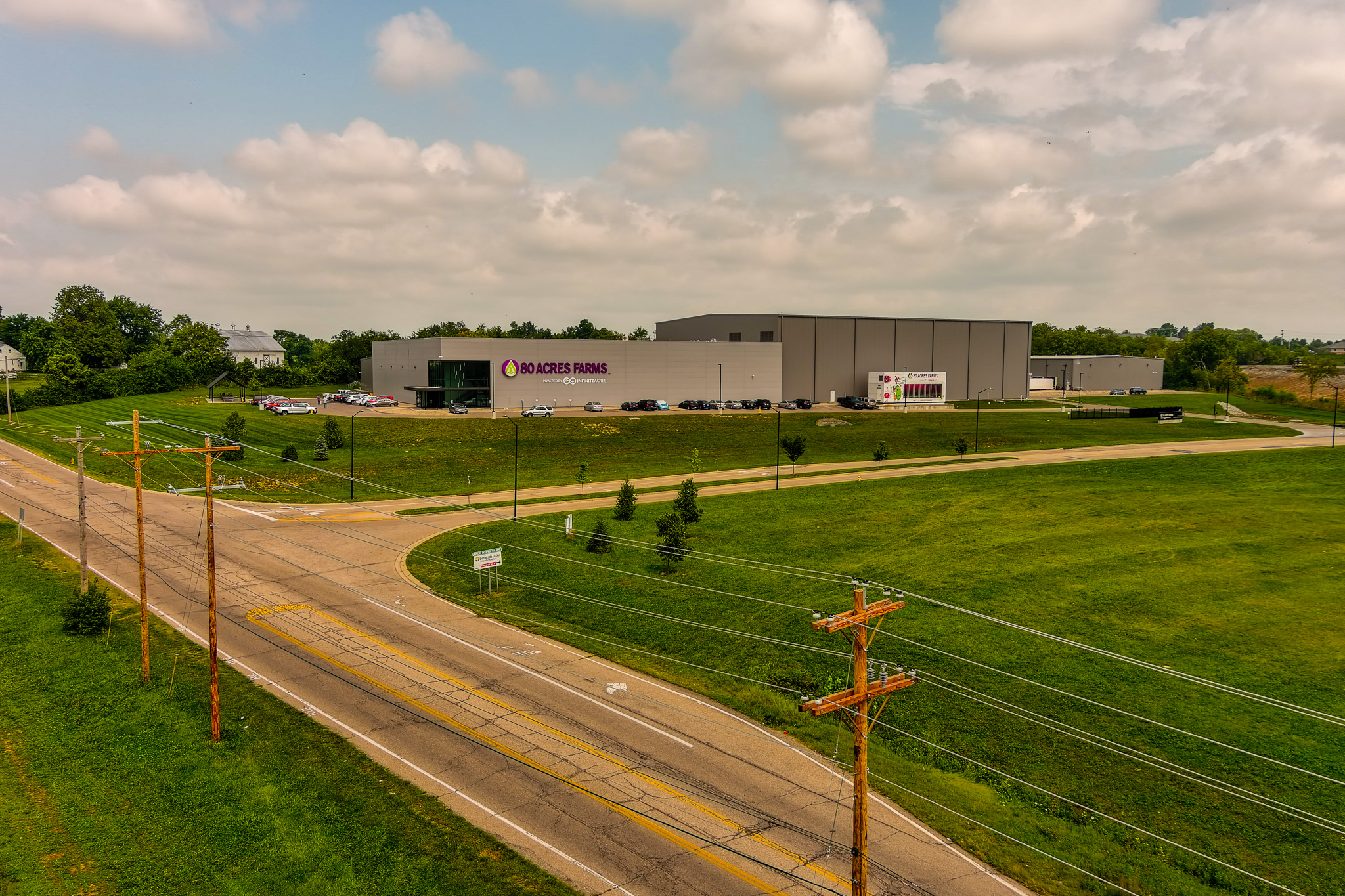 Warehouse beside a road, surrounded by green fields under a cloudy sky.