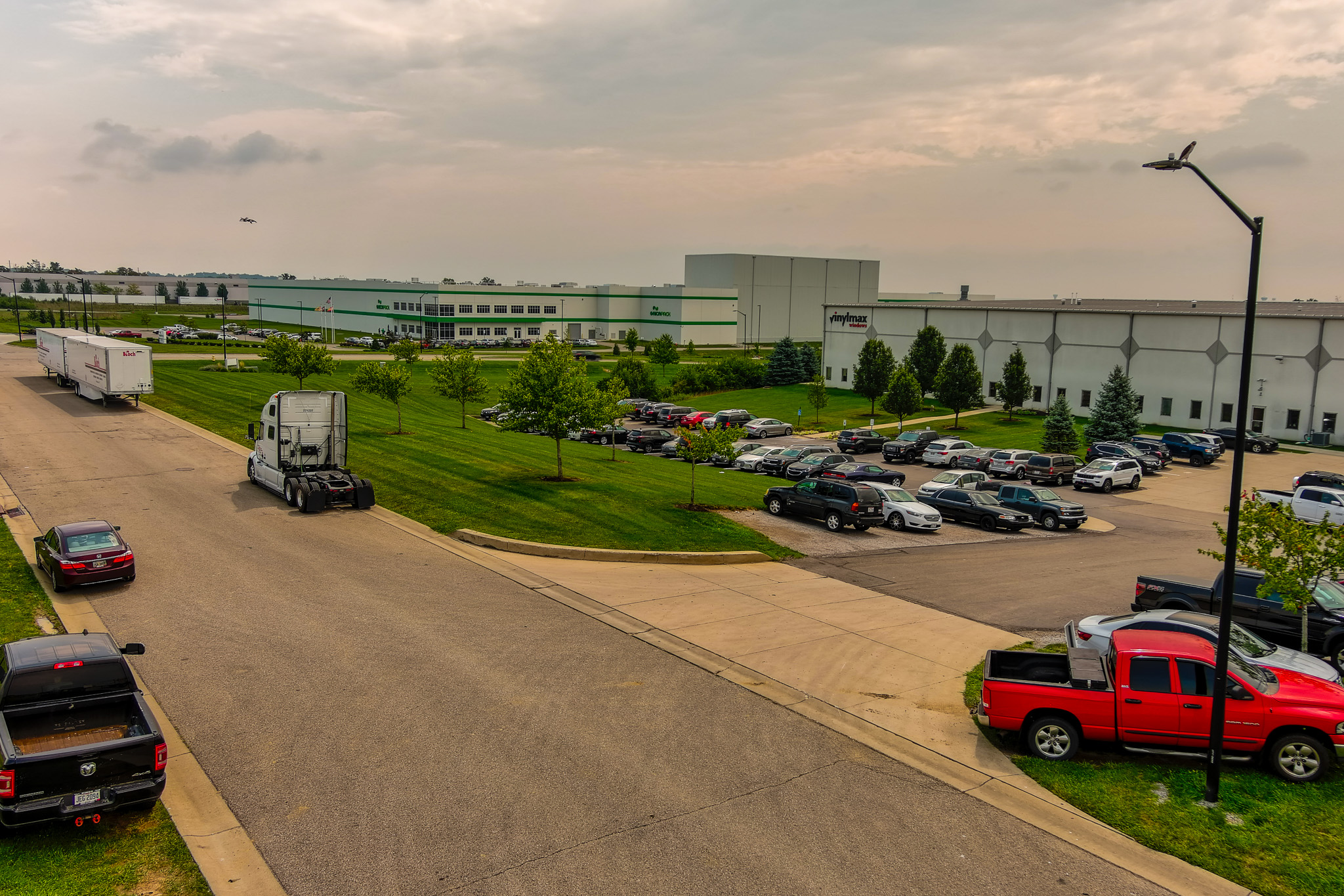 Industrial park with warehouses, parked trucks, and cars under a cloudy sky.