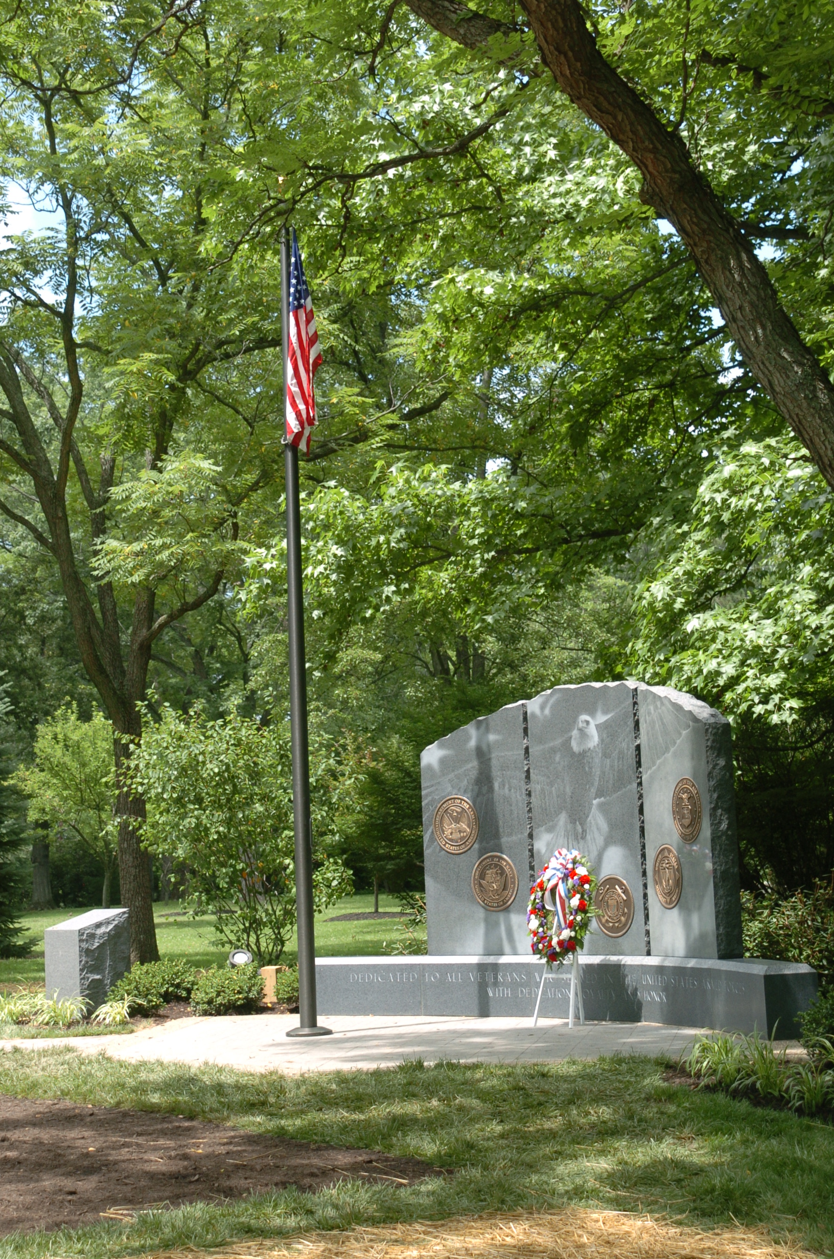 Memorial with wreath, trees, and flagpole in a green park setting.