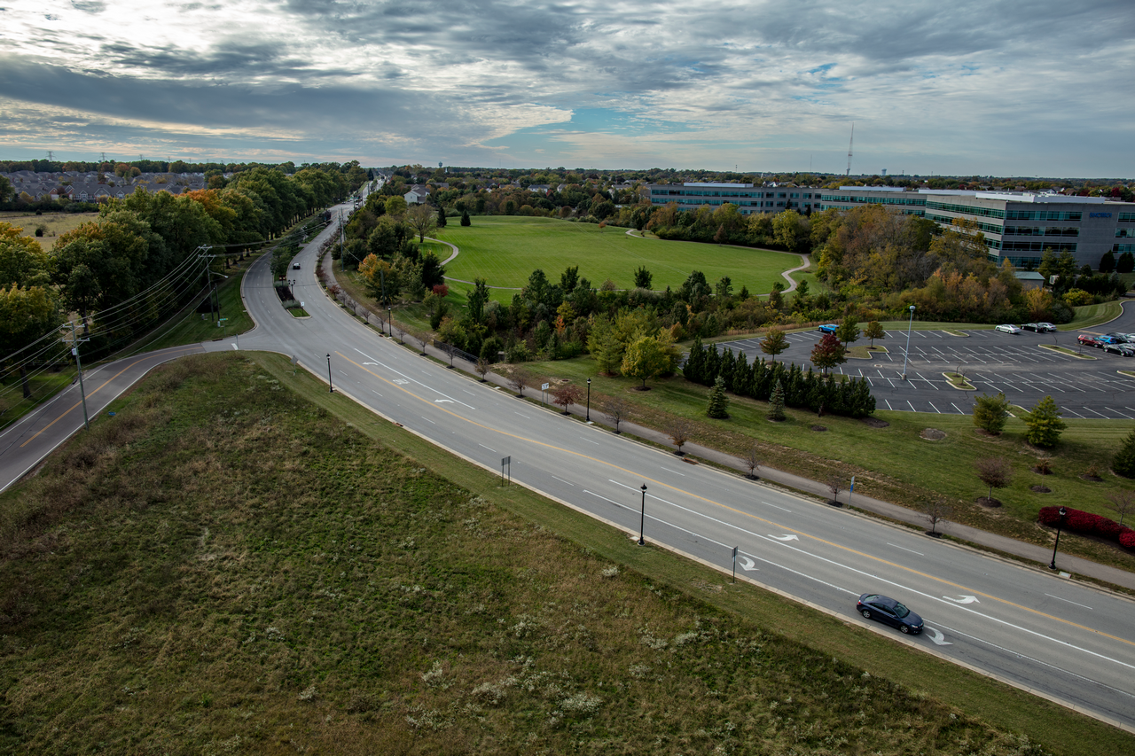 Winding road through grassy landscape, with cars and distant buildings under a cloudy sky.