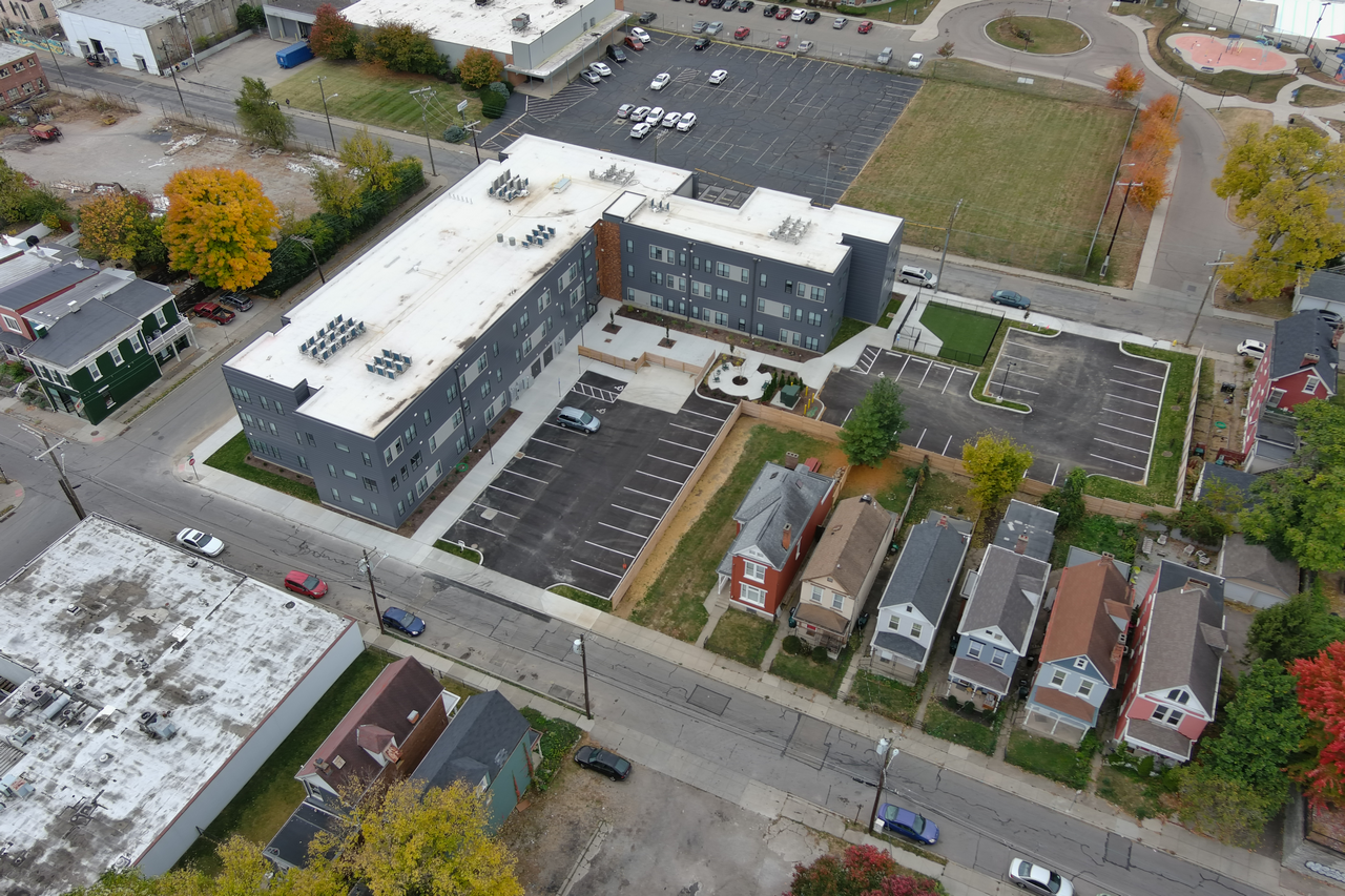 Aerial view of a modern building beside a row of colorful houses, surrounded by trees.