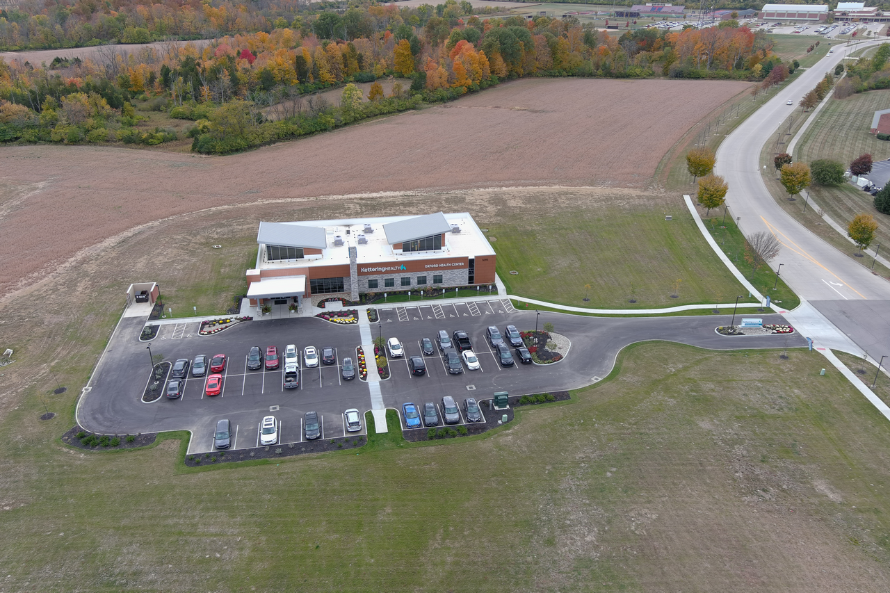 Aerial view of a building with a parking lot, surrounded by fields and a road nearby.