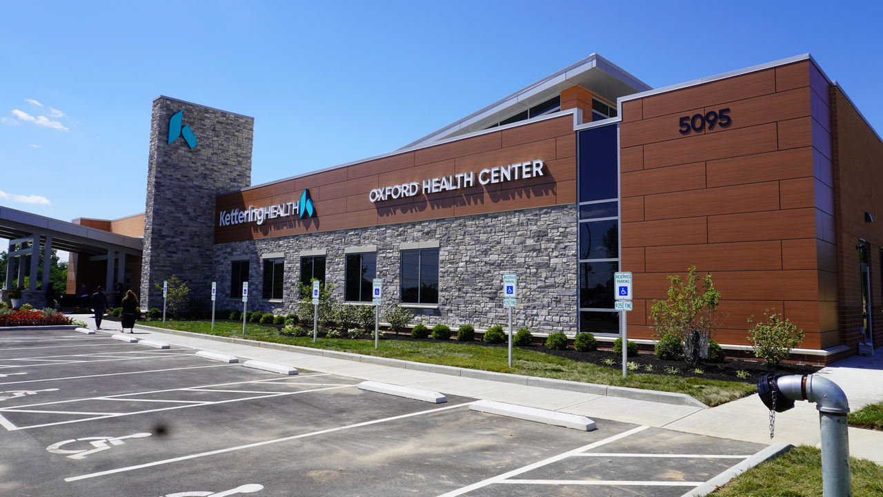Modern health center building with stone and wood facade, parking lot in foreground.