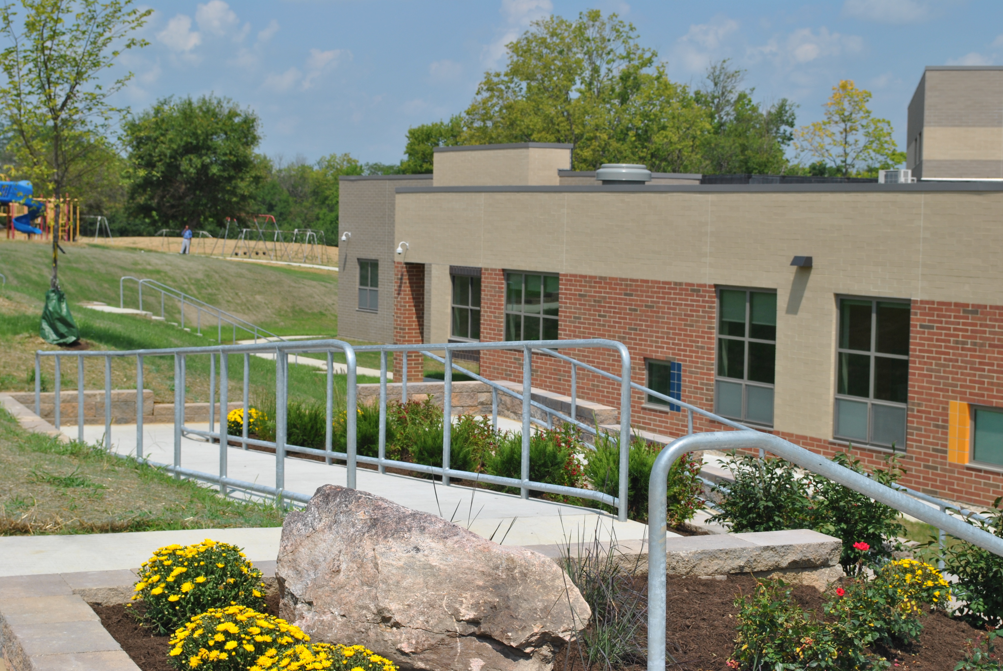 Accessible ramp with metal railing outside a brick building, surrounded by greenery.