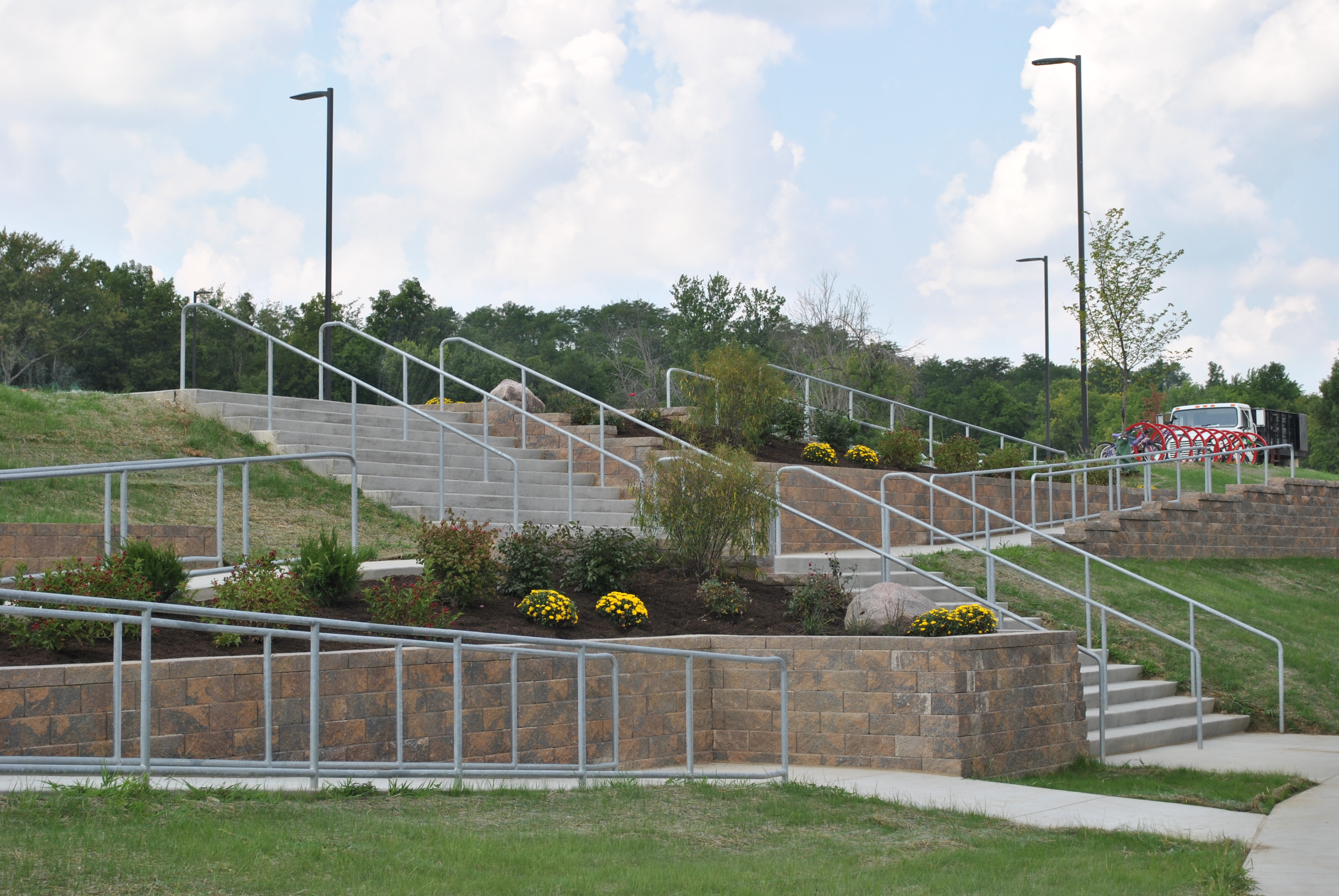 Outdoor stairway between landscaped terraces with railings and lampposts.