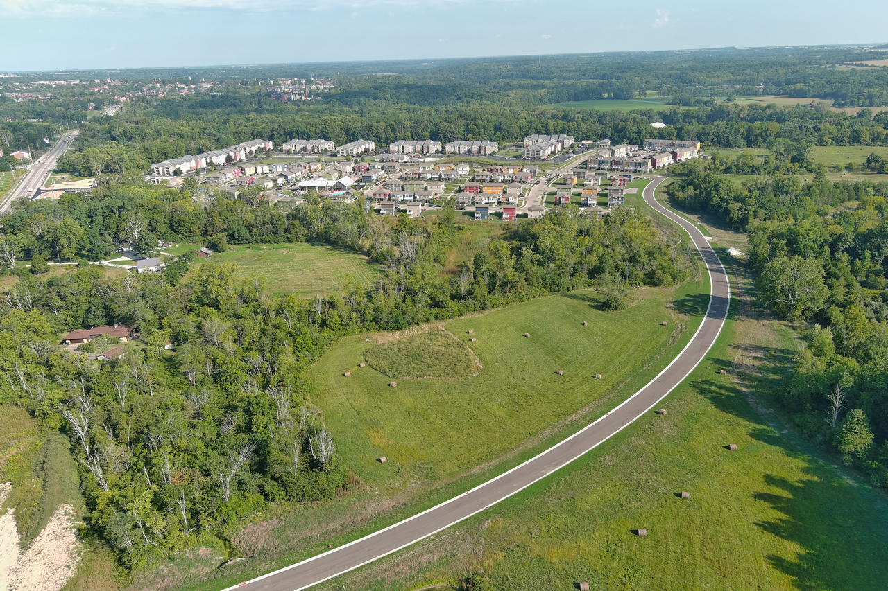 Aerial view of a road curving through fields and trees, with houses in the distance.