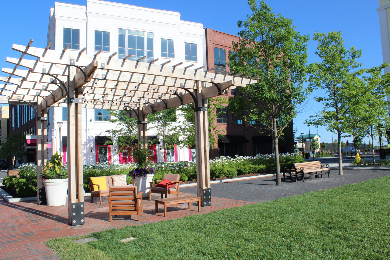 Wooden pergola with seating on a sunny day, flanked by trees and a modern building.