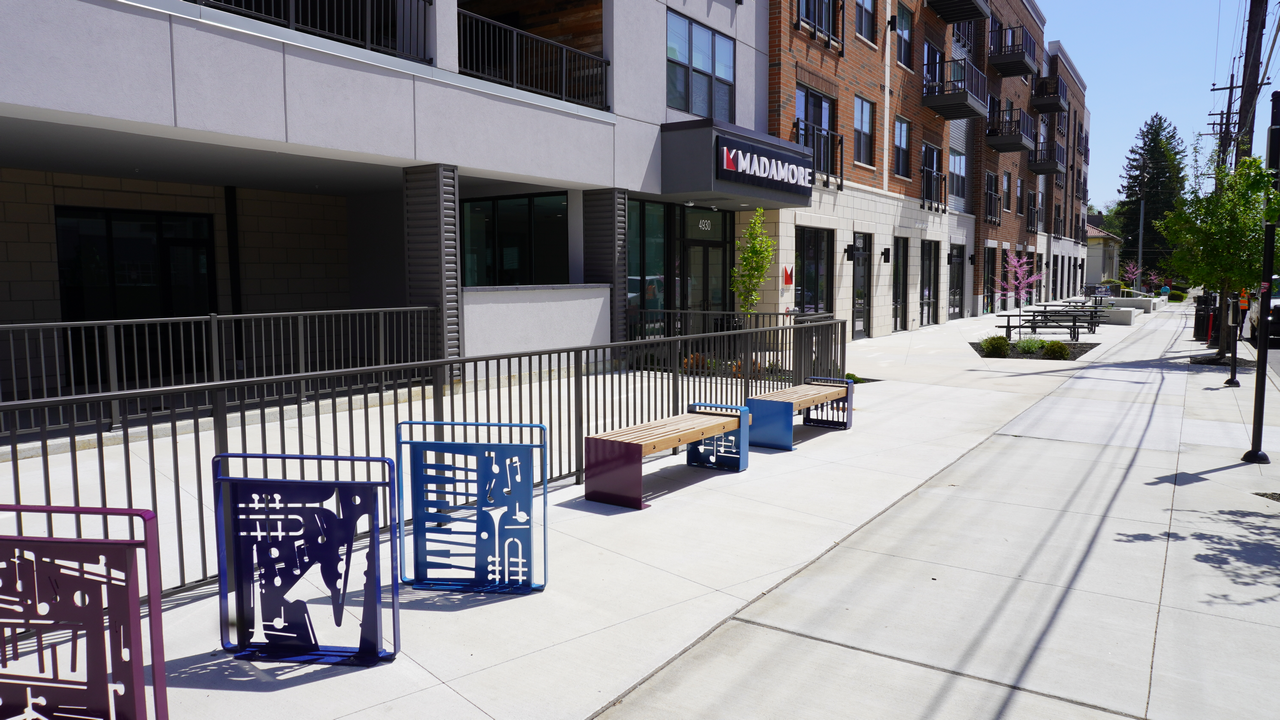 Modern apartment building with wide sidewalk, colorful benches, and trees lining the street.