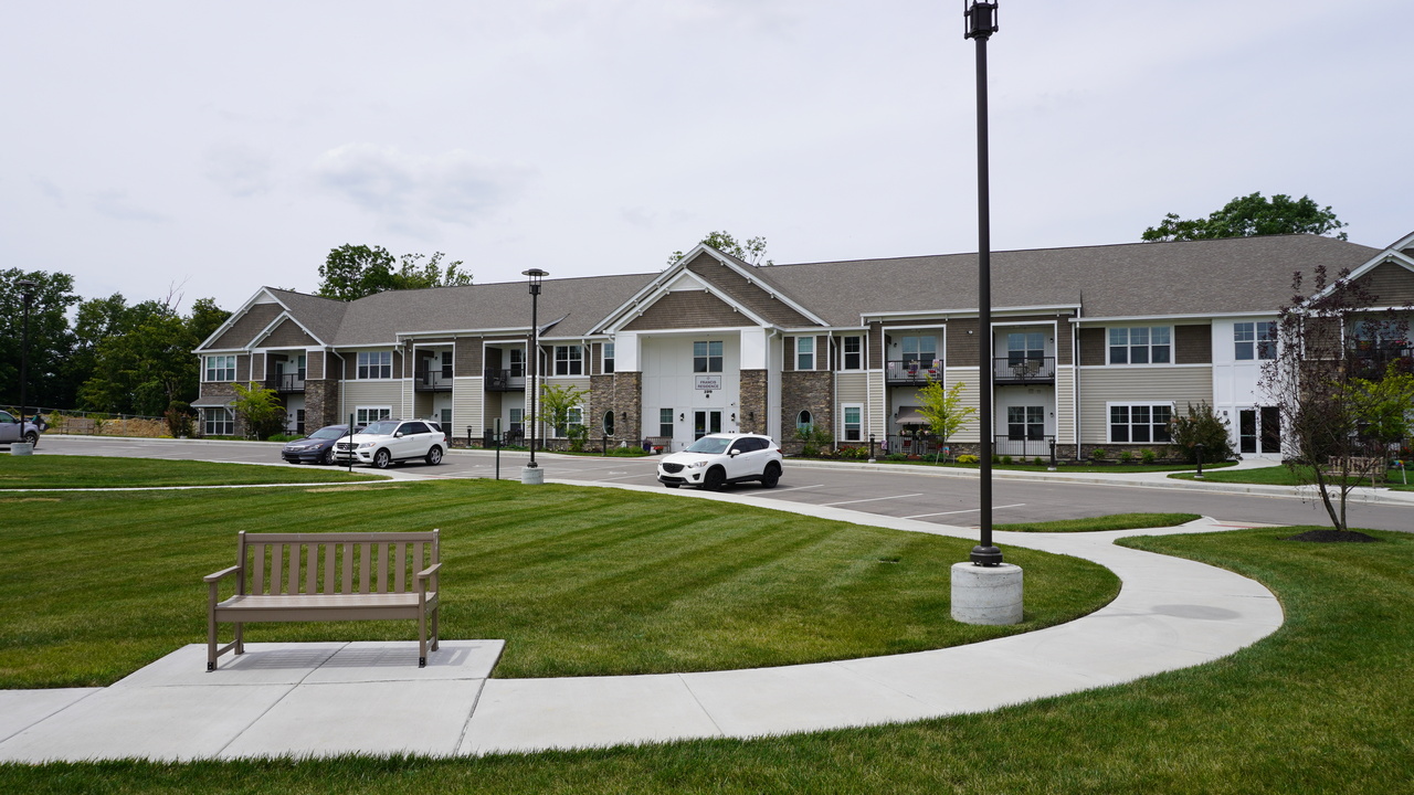 Apartment complex with parked cars, green lawn, and a bench. Overcast sky.