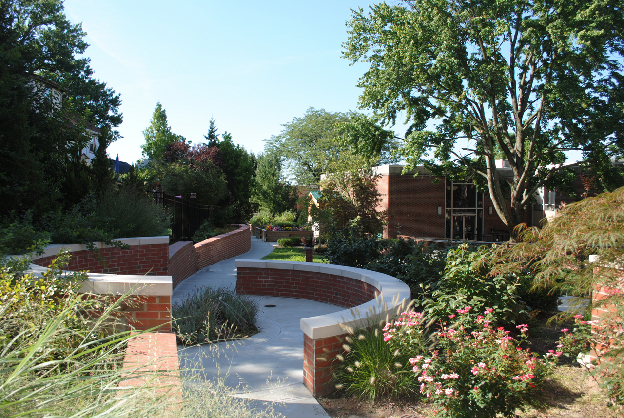 Curved brick walkway through garden with trees and shrubs.