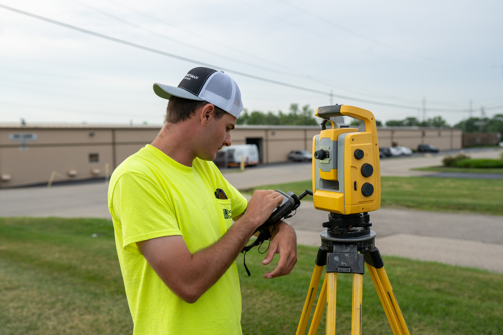 Man in yellow shirt adjusting survey equipment on tripod outdoors.