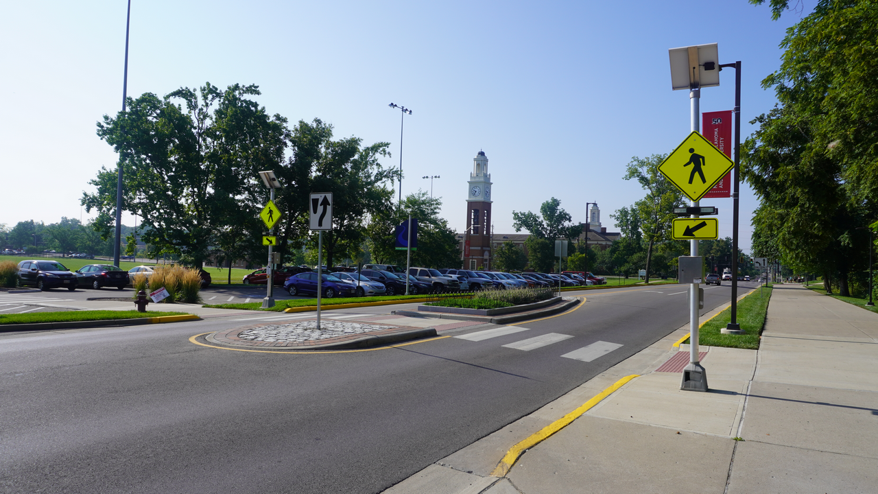 Campus road with crosswalk, parking lot, and distant clock tower under clear blue sky.