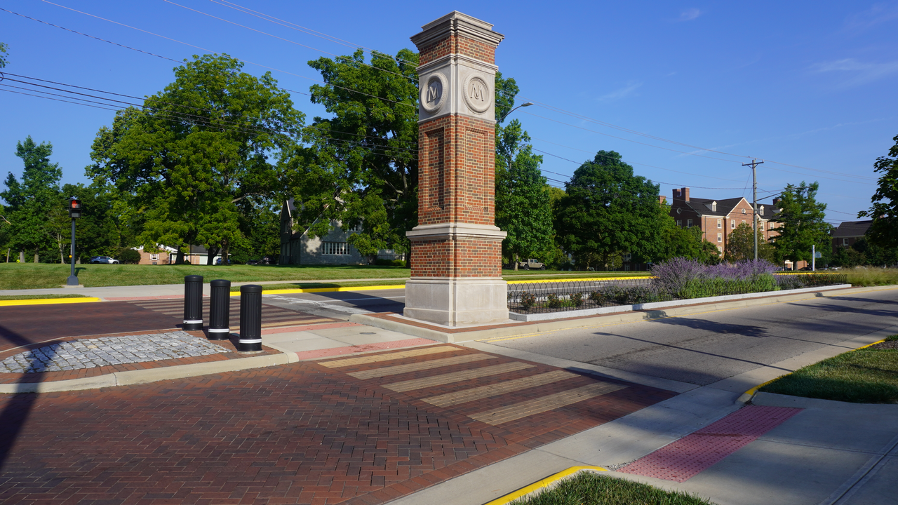 Brick clock tower on a sunny street corner with trees and a blue sky.