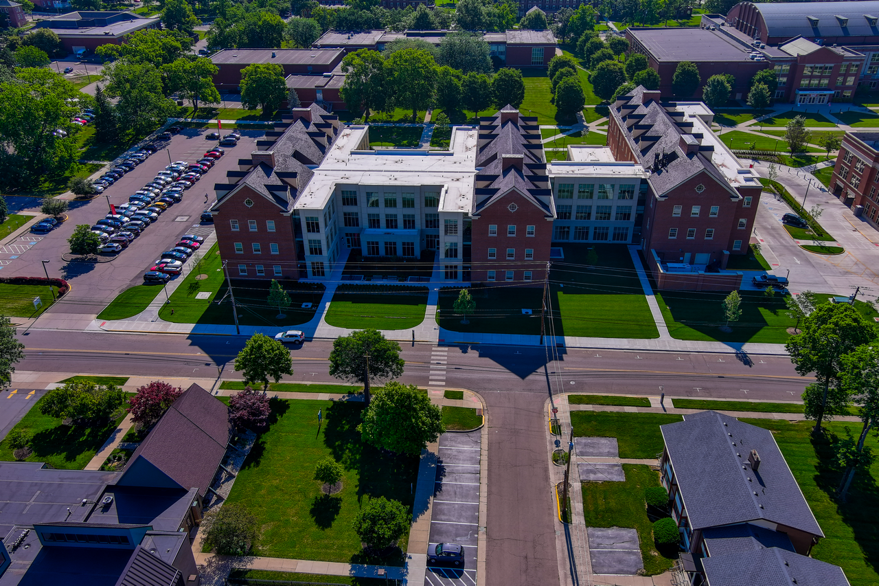 Aerial view of a symmetrical brick building complex with greenery and parked cars.