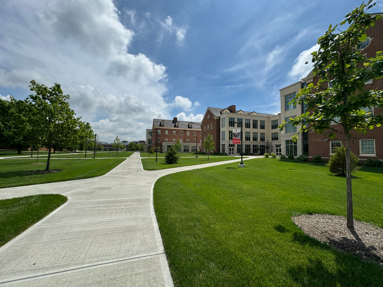 Campus path with green lawns, trees, and brick buildings under a cloudy sky.