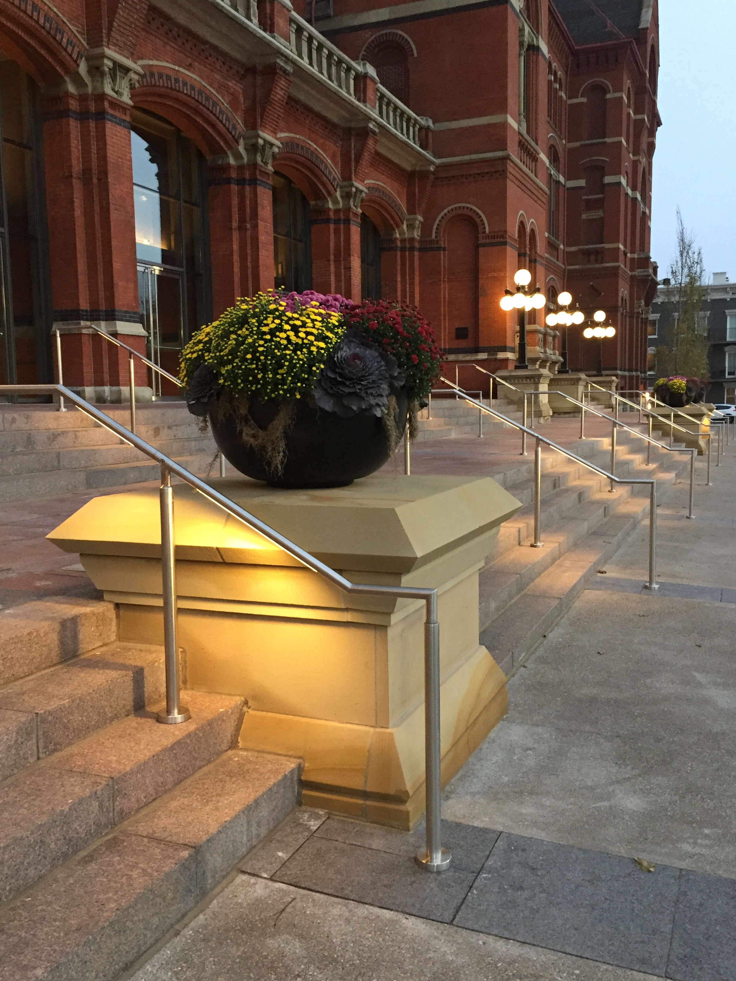 Large potted flowers on a lit stone pedestal near a red brick building entrance.