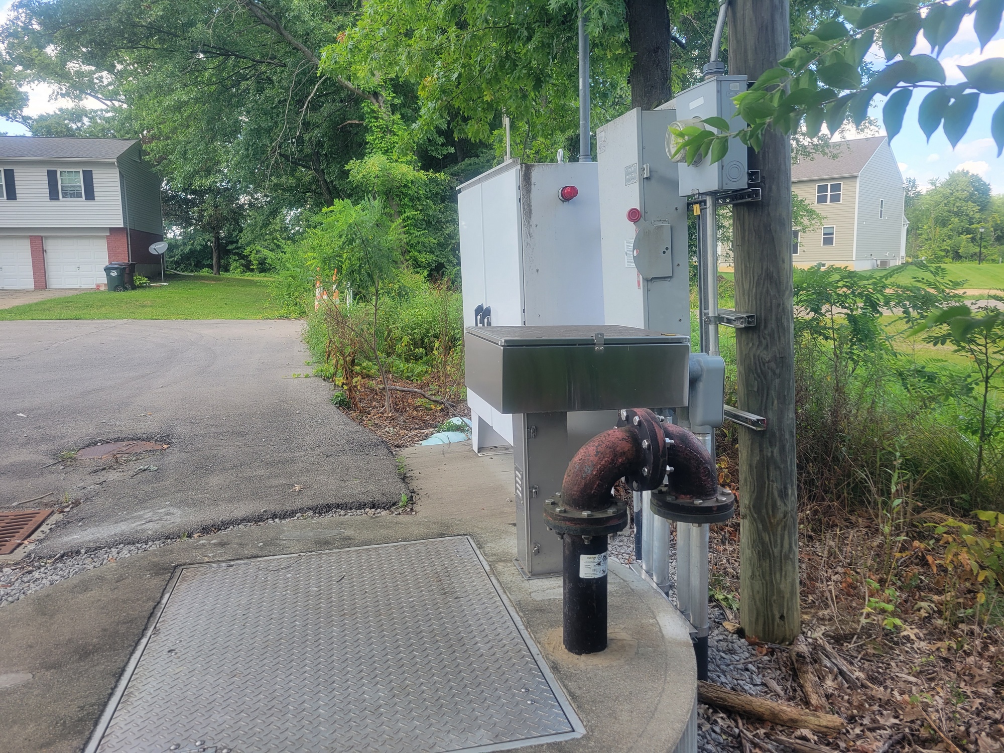 Electrical box and pipes near a road with houses and trees in the background.