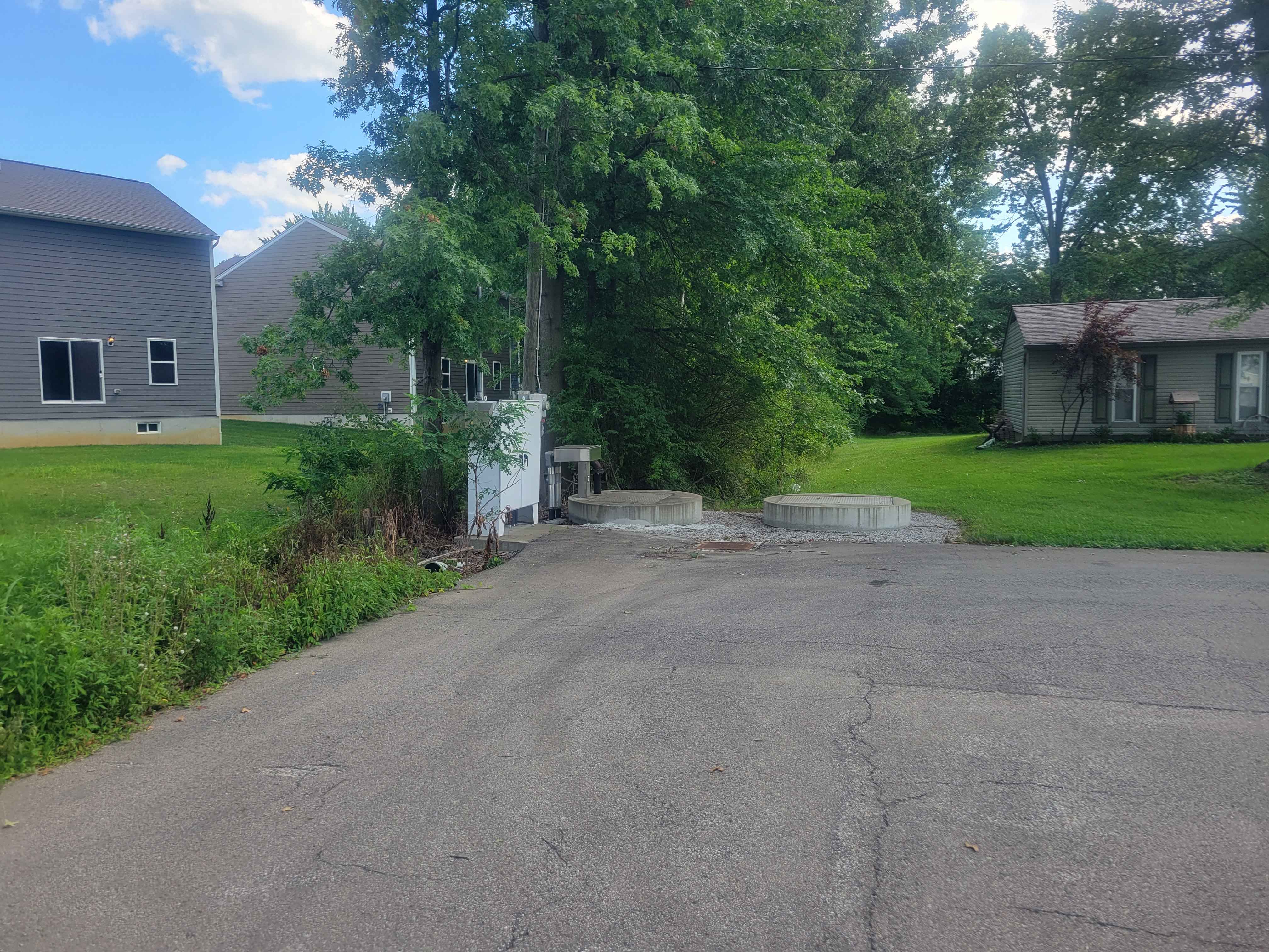 Road ending at grass with trees and houses nearby under a blue sky.
