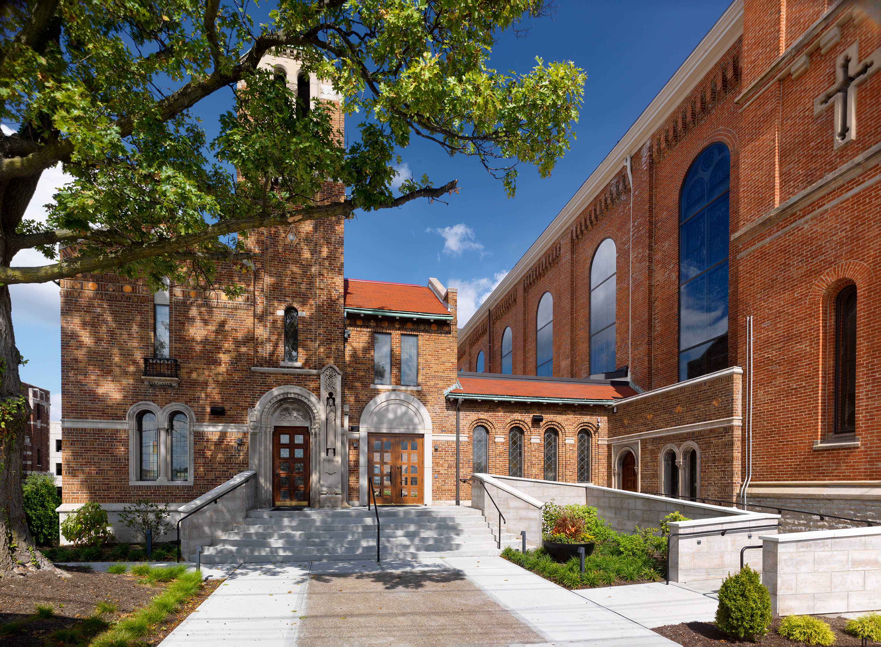 Historic brick church with arched windows, sunny day, large tree in foreground.