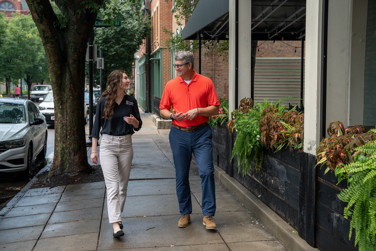 Man and woman walking on a sidewalk, talking and smiling.