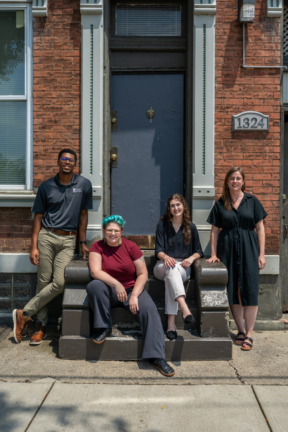 Four people smiling outside a brick building, some sitting, some standing.