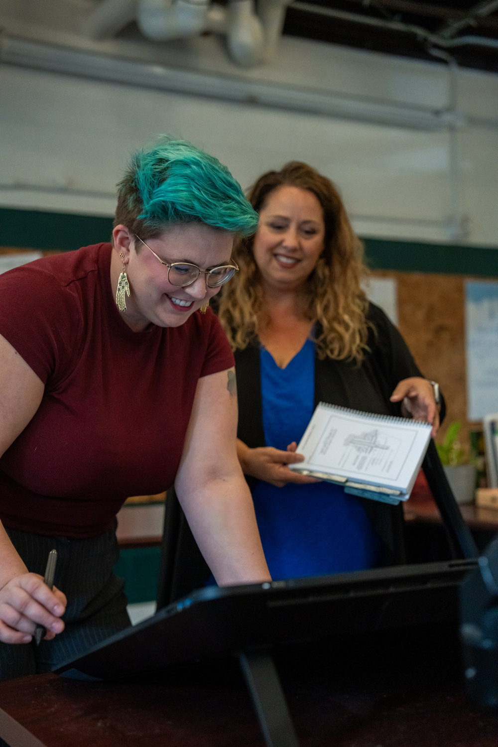 Two women smiling while working at a desk, one writing, the other holding a notebook.