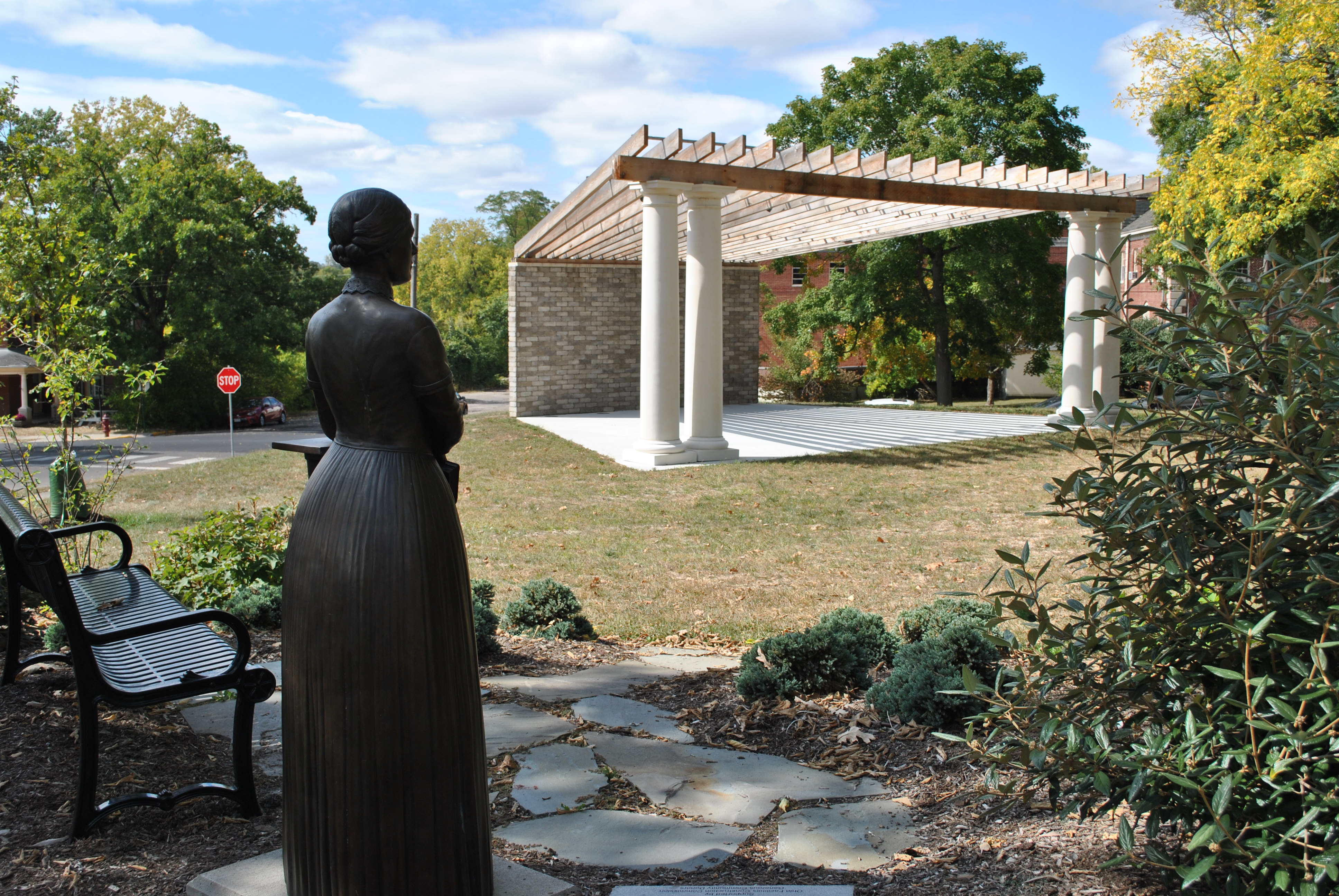 Stone statue of a woman near benches facing a gazebo in a park.